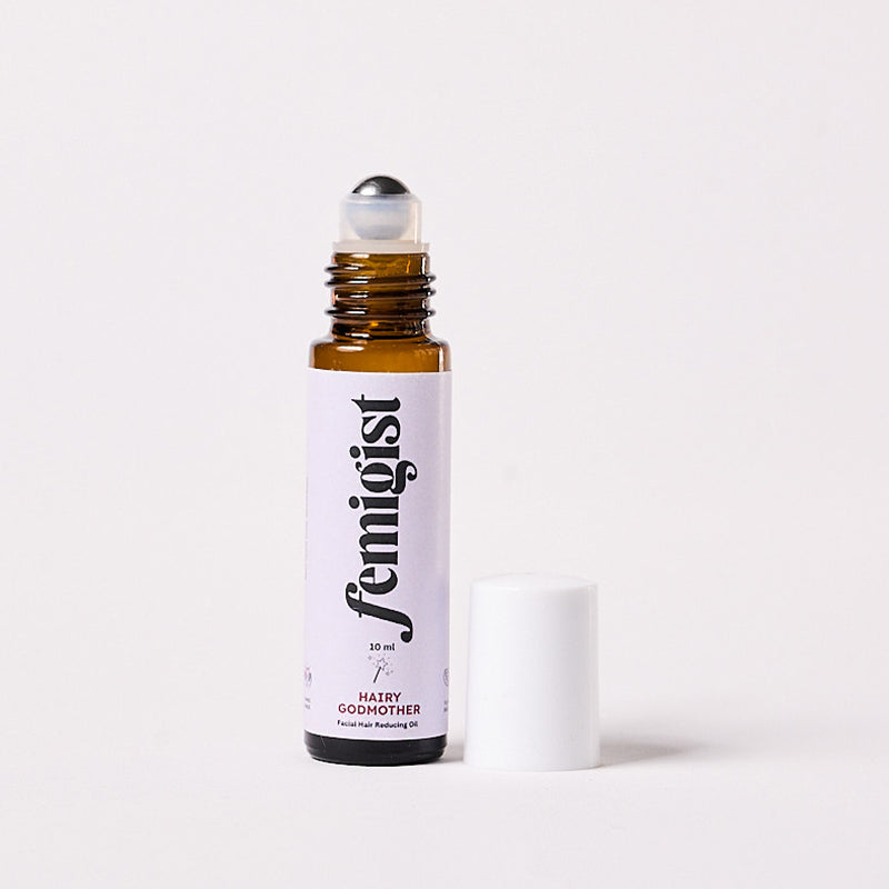 10ml roller bottle of Hairy Godmother facial hair reducing oil by Femigist, with the cap removed and the roll-on top visible, set against a white background.
